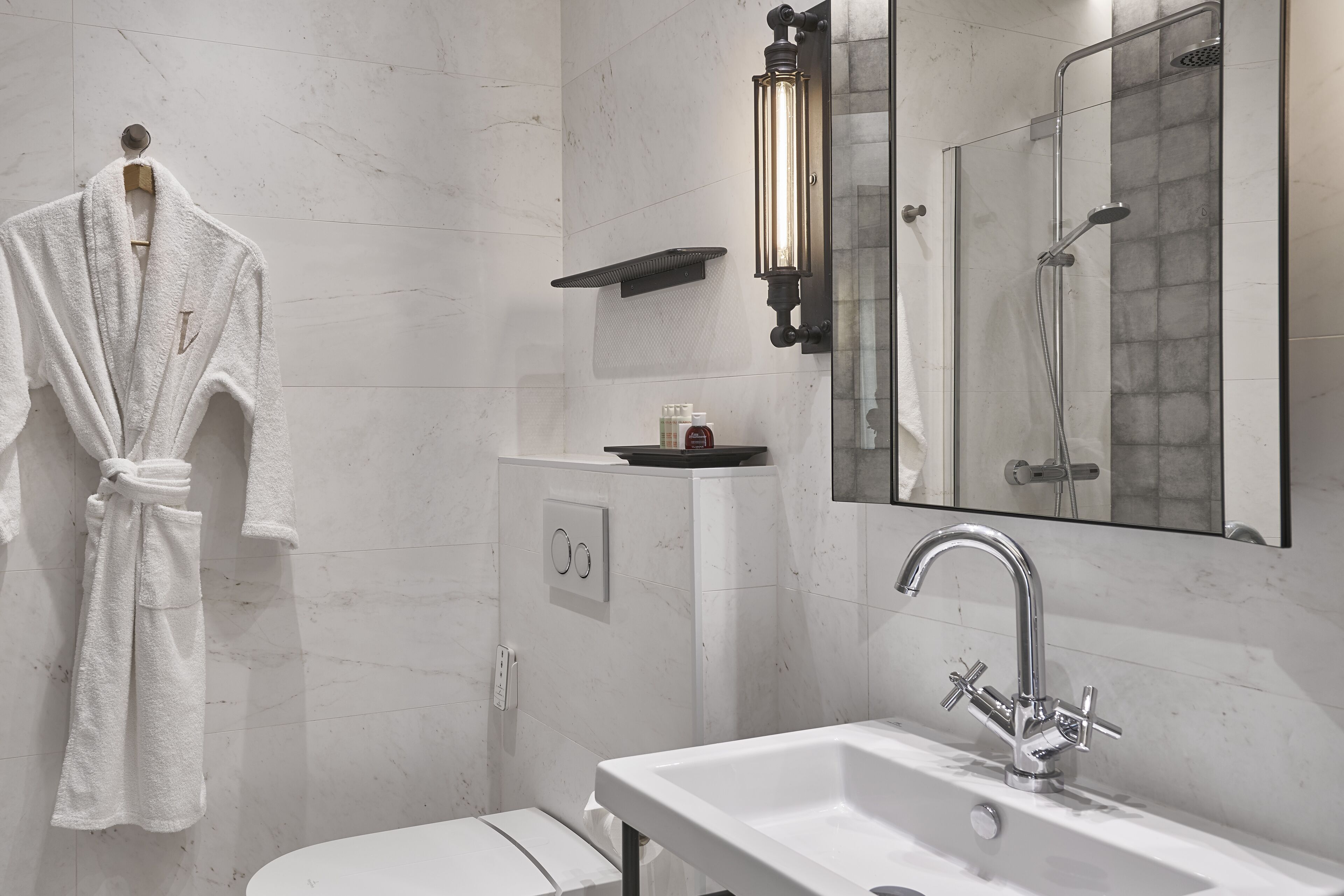 Un baño moderno con un lavamanos blanco, cabina de ducha de vidrio, una bata colgada en la pared y un aplique en la pared.