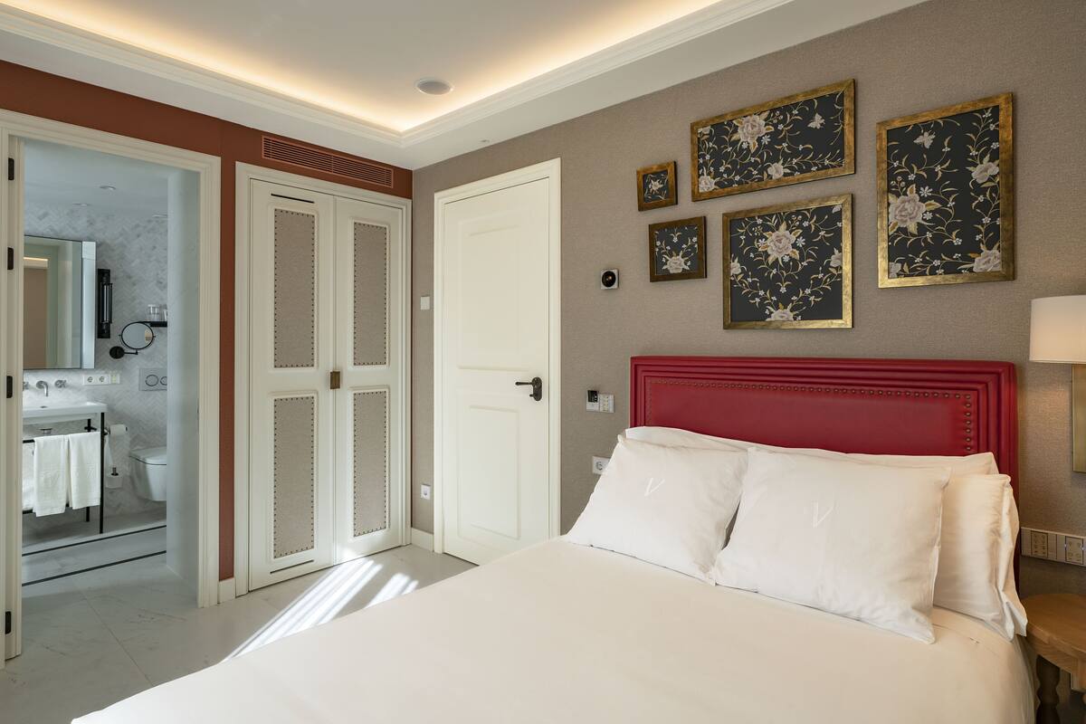 A hotel room with a bed, a red headboard, a white door, and a bathroom visible through an open door.