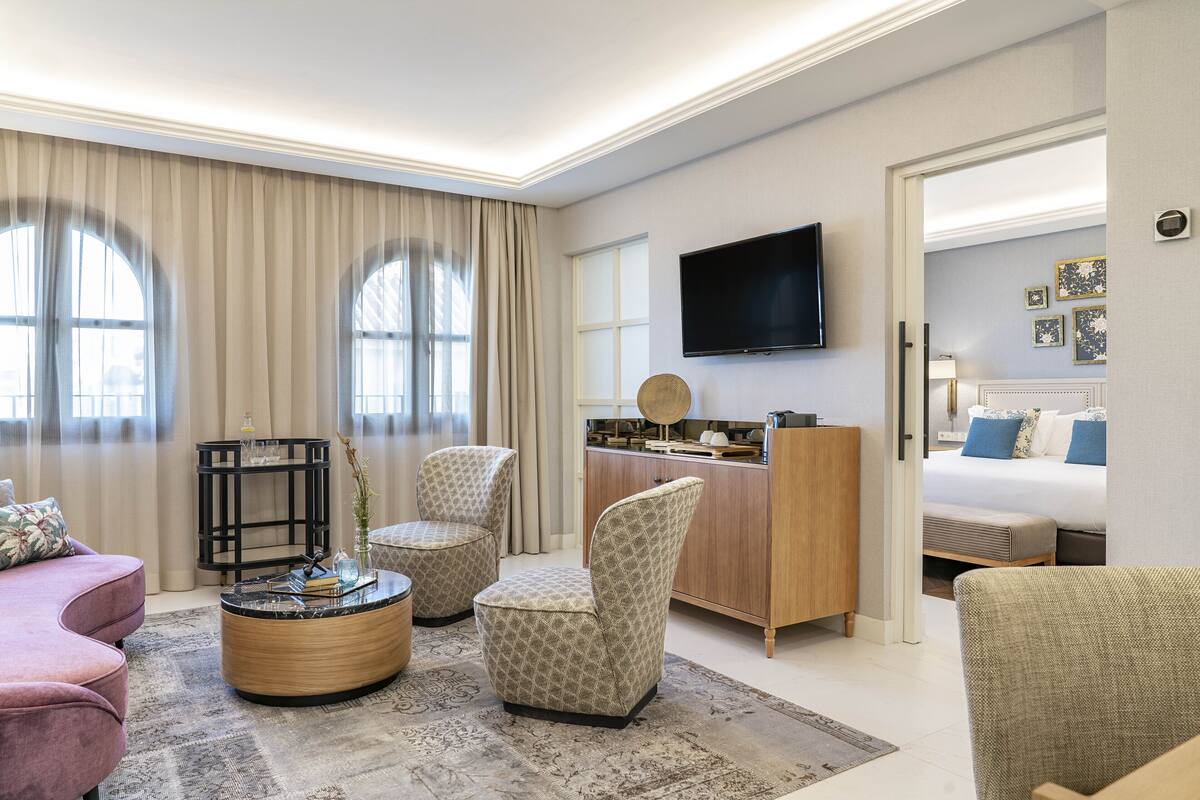 A modern hotel room with a sofa, two armchairs, a coffee table, a desk, a flat-screen TV, and a bed.