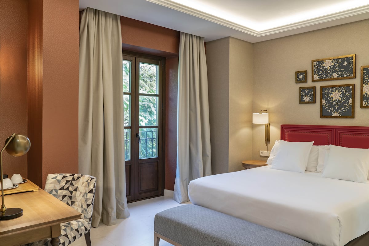 A hotel room with a bed, a desk with a lamp, a chair, and a window with curtains.