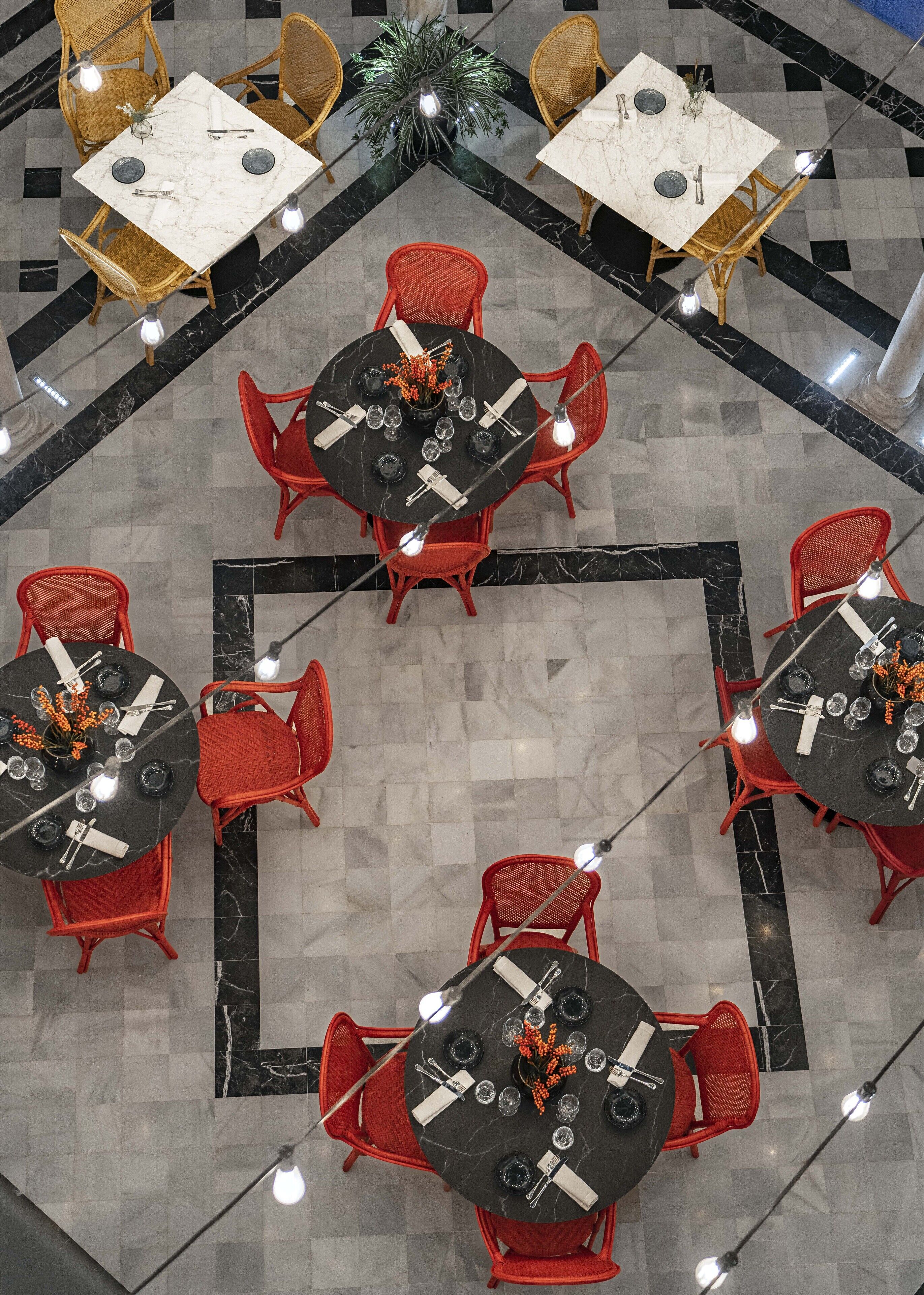 A hotel lobby with multiple dining tables, some with red chairs and others with wicker chairs, arranged in a circular pattern.