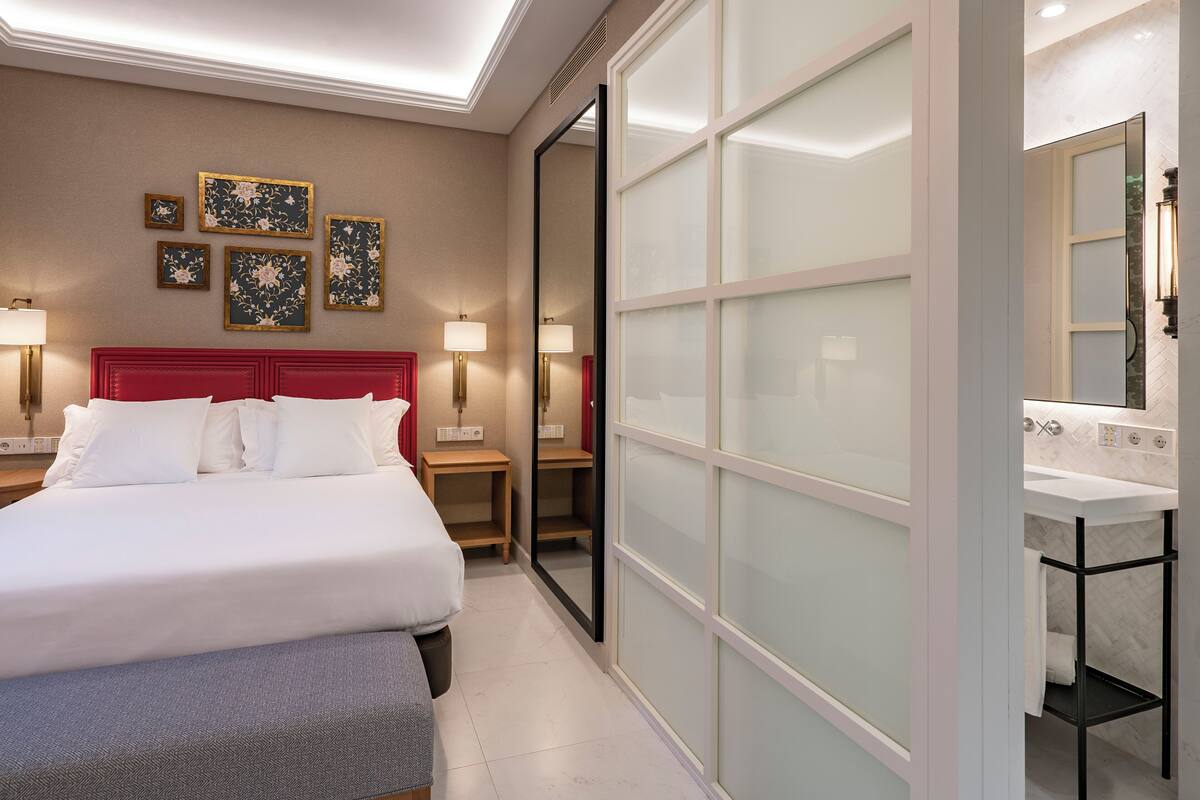A hotel room with a large bed, two bedside tables with lamps, a wardrobe with frosted glass doors, and a bathroom with a sink and mirror.