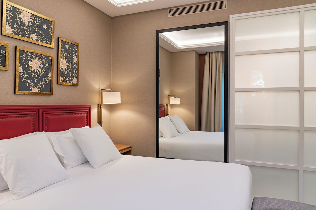 A hotel room with a large bed, a red headboard, a nightstand with a lamp, and three framed floral artworks on the wall.