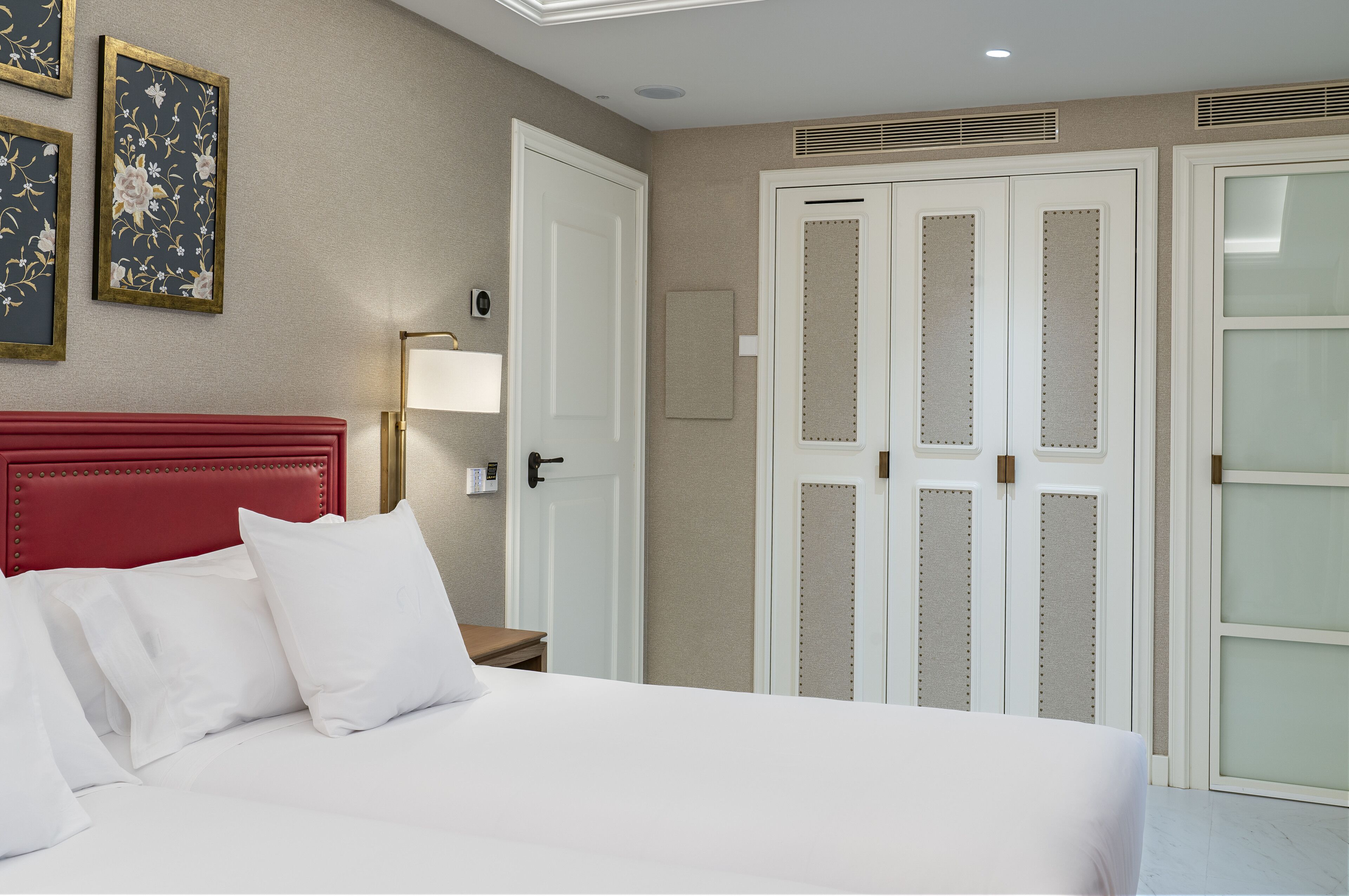 Une chambre d’hôtel avec une tête de lit rouge, du linge de lit blanc, une lampe de chevet et une armoire aux portes en verre dépoli.