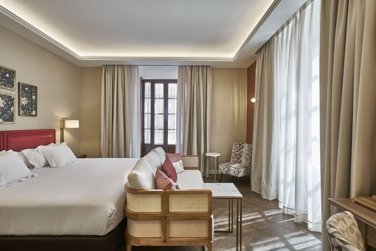 A hotel room with a large bed, a sofa, a small table, and a chair. There are curtains on the windows and a lamp on the table.