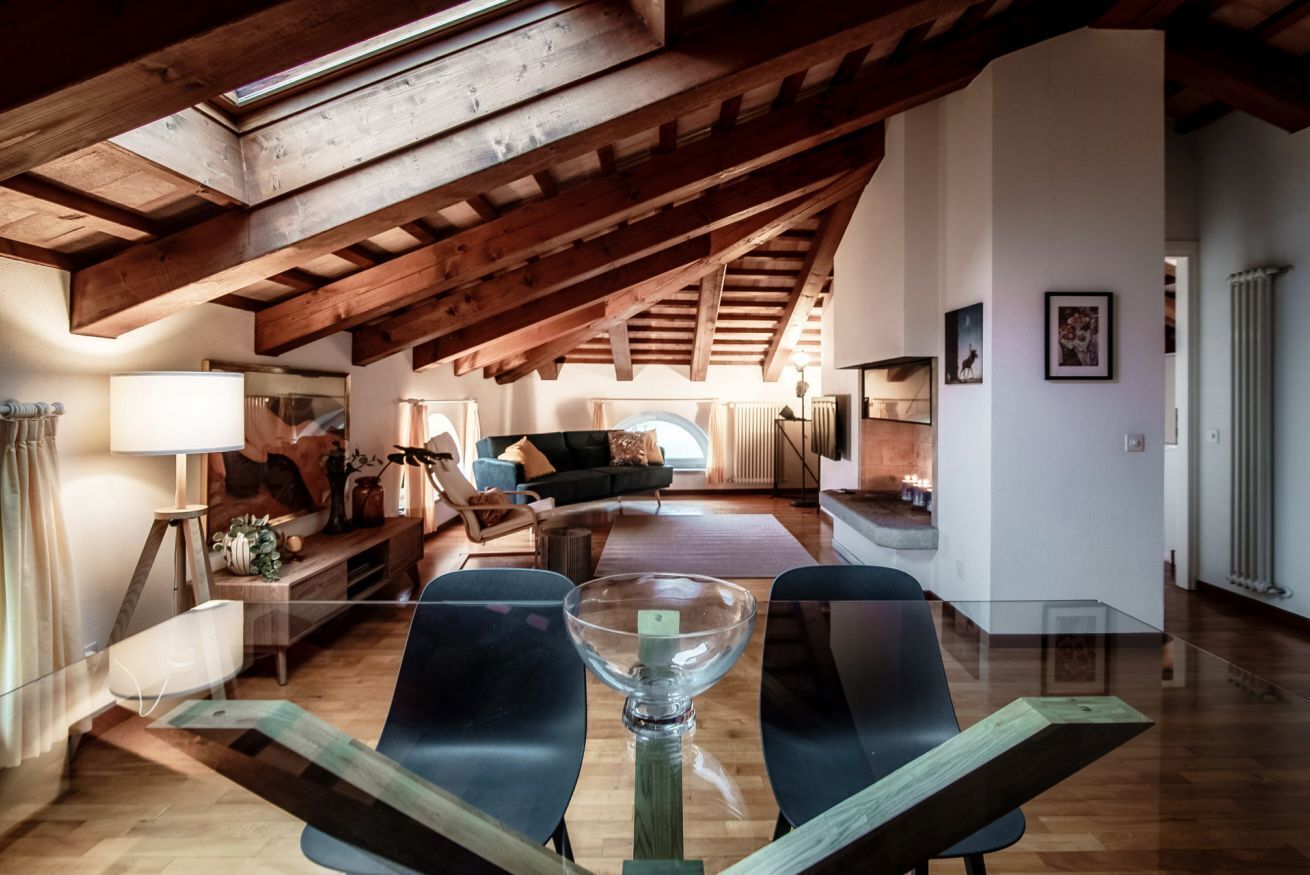 A spacious loft with exposed wooden beams, a glass dining table, and a cozy living area with a sofa and fireplace.