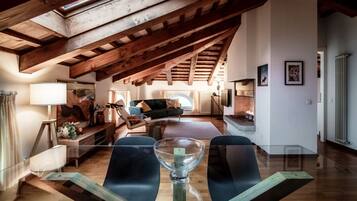 A spacious loft with exposed wooden beams, a glass dining table, and a cozy living area with a sofa and fireplace.