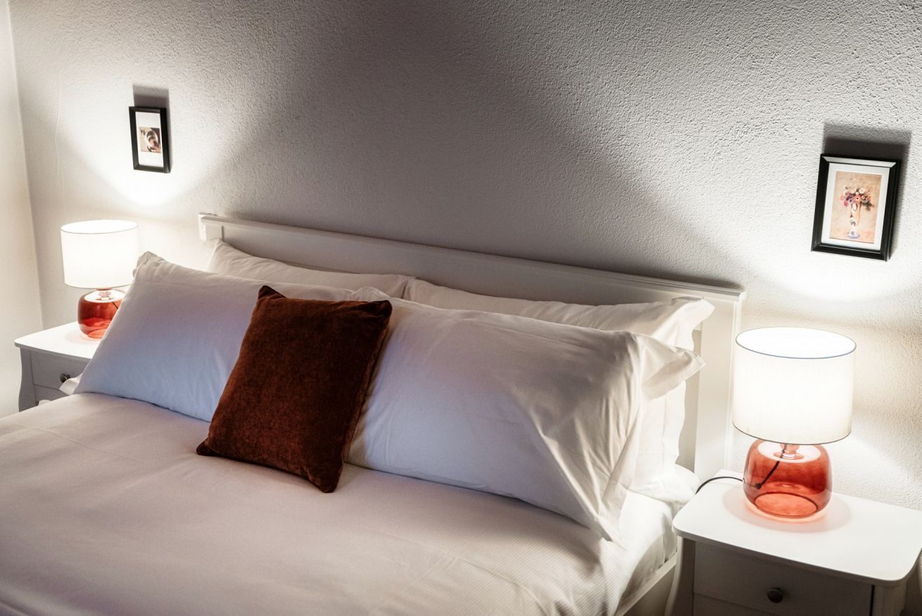 Ein ordentlich bezogenes Bett mit weißen Leinentextilien und einem braunen Zierkissen, flankiert von zwei Nachttischen mit Lampen.
