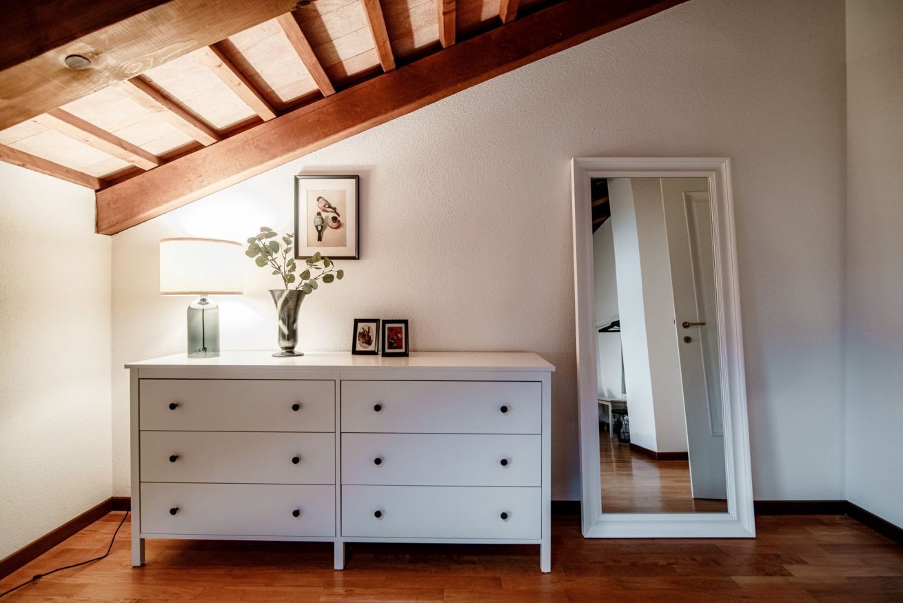 A white dresser with drawers, a vase with green leaves, and two framed pictures. There is a lamp with a white shade and a mirror on the wall.