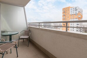 A balcony with a glass table and two wicker chairs, overlooking a cityscape with modern buildings.