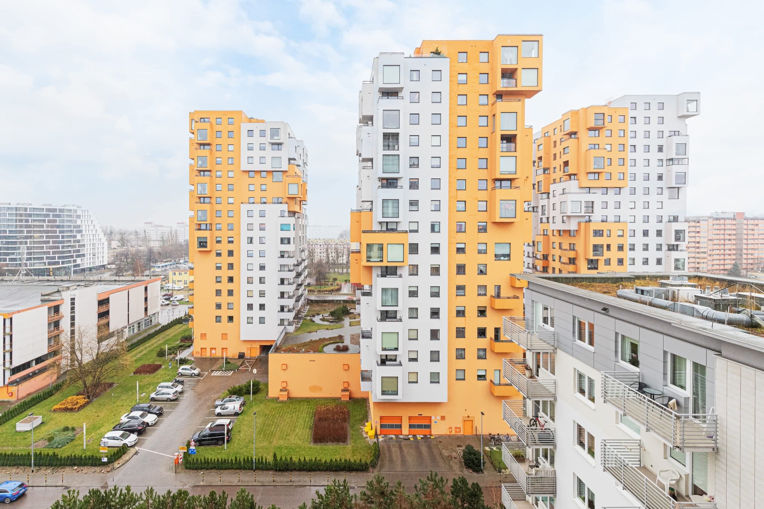 A high-rise residential building with multiple balconies, a parking lot, and a small green area.