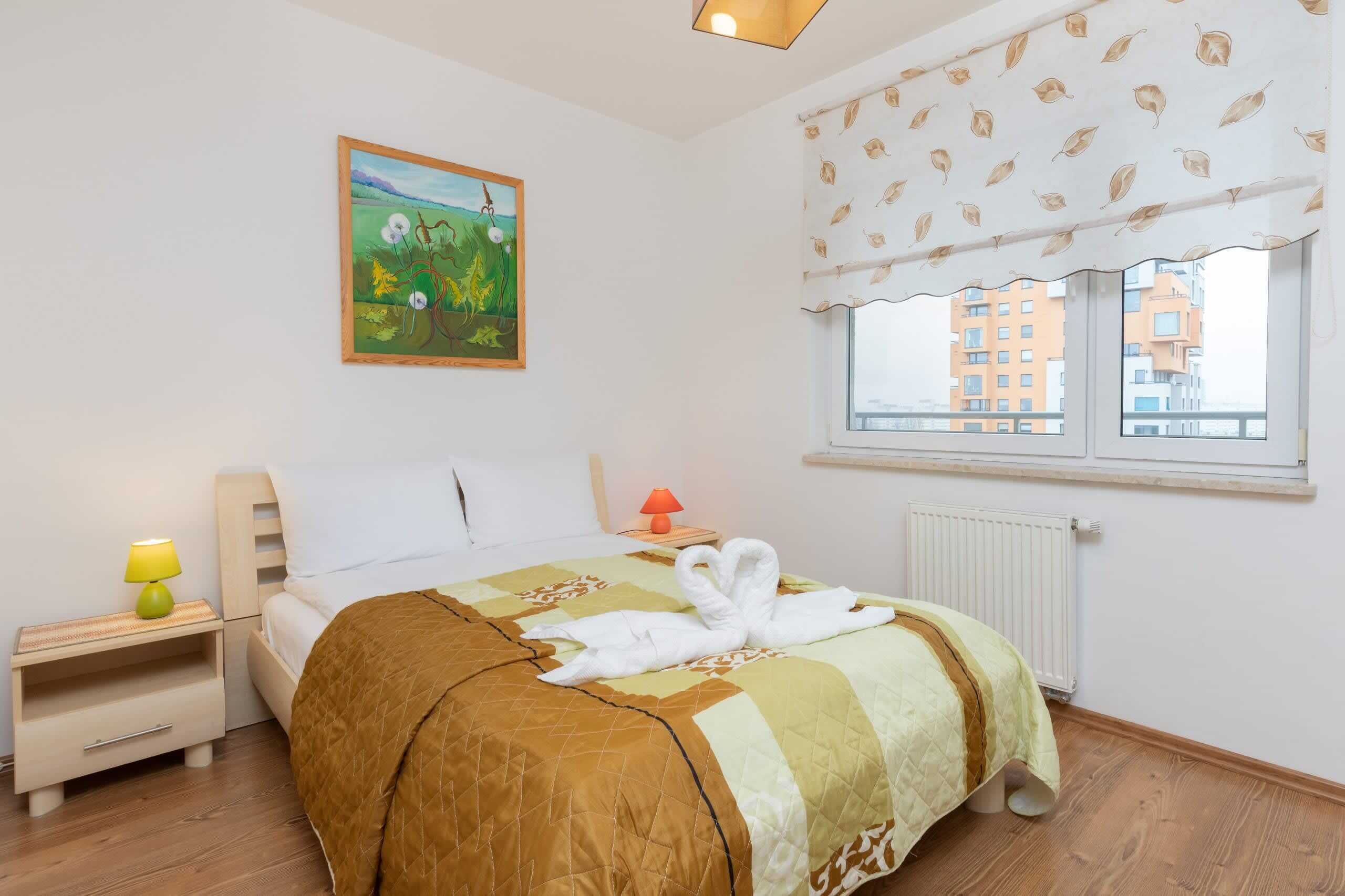 A neatly made bed with a quilt, a bedside lamp, a painting on the wall, and a window with a view of buildings.