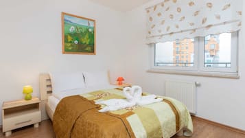 A neatly made bed with a quilt, a bedside lamp, a painting on the wall, and a window with a view of buildings.