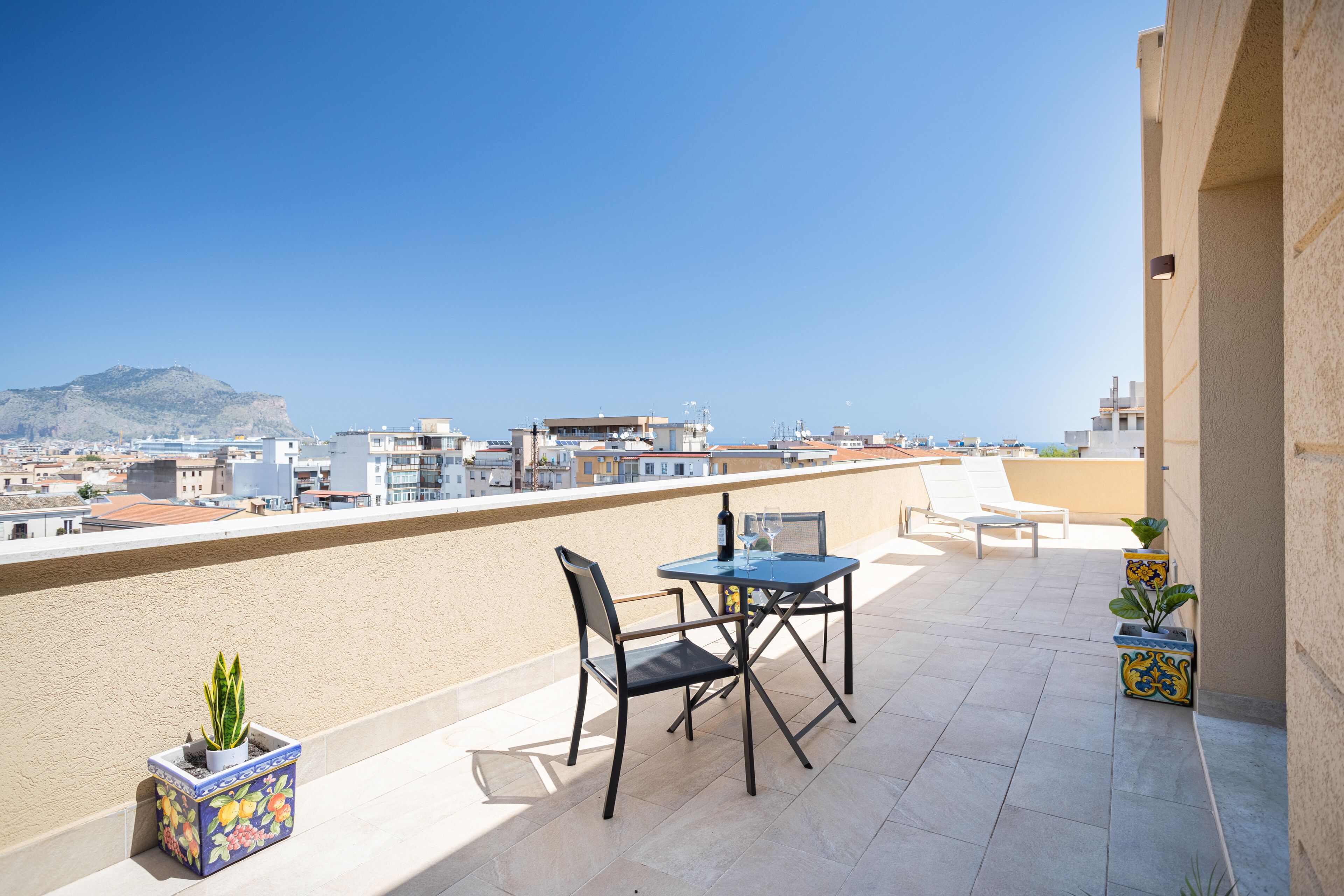 Una terraza en la azotea con mesa y sillas, plantas en macetas y vistas a la ciudad y a las montañas.