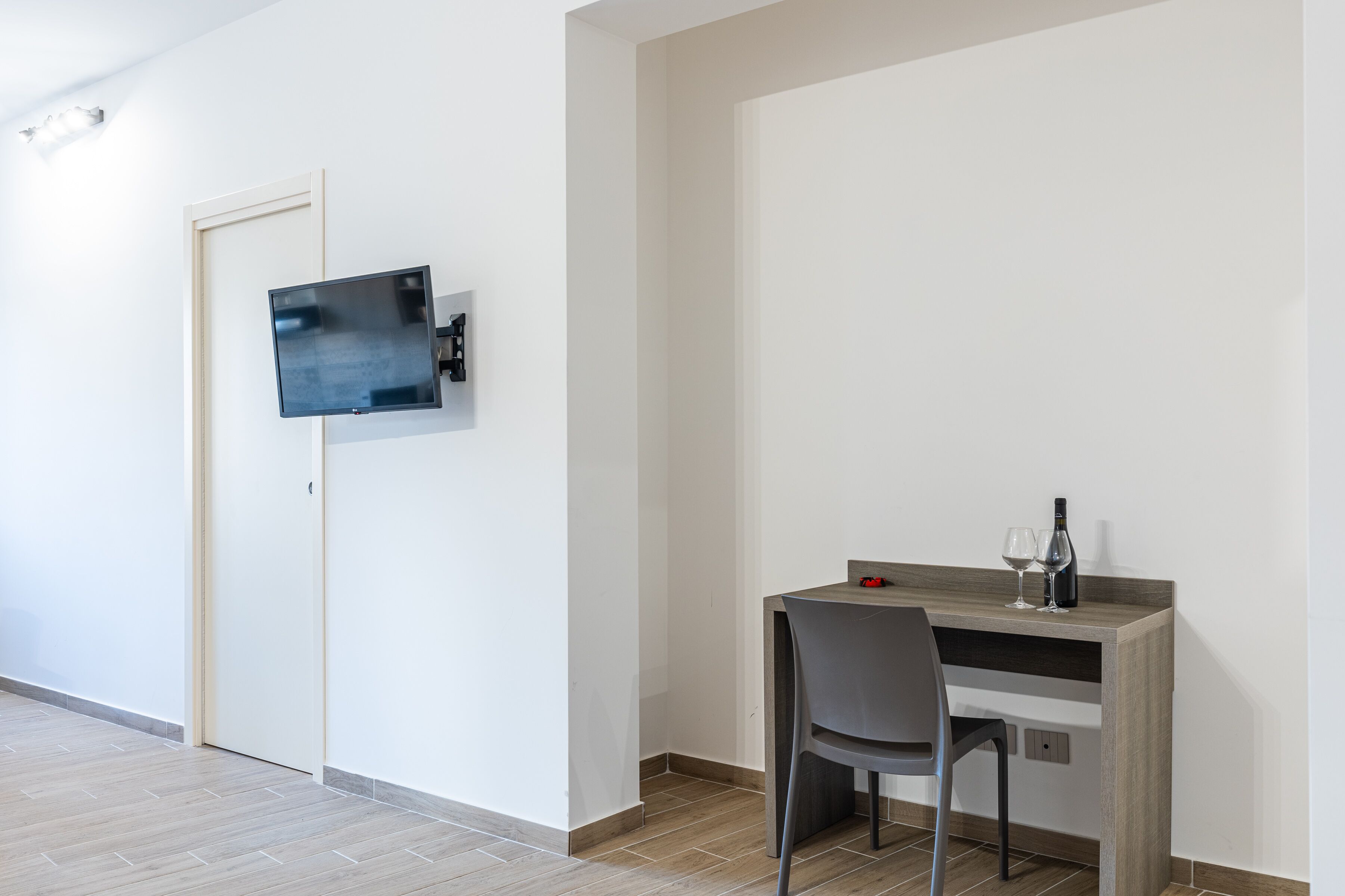 A modern room with a flat-screen TV mounted on the wall, a wooden desk with a chair, and a wine bottle and glass on the desk.