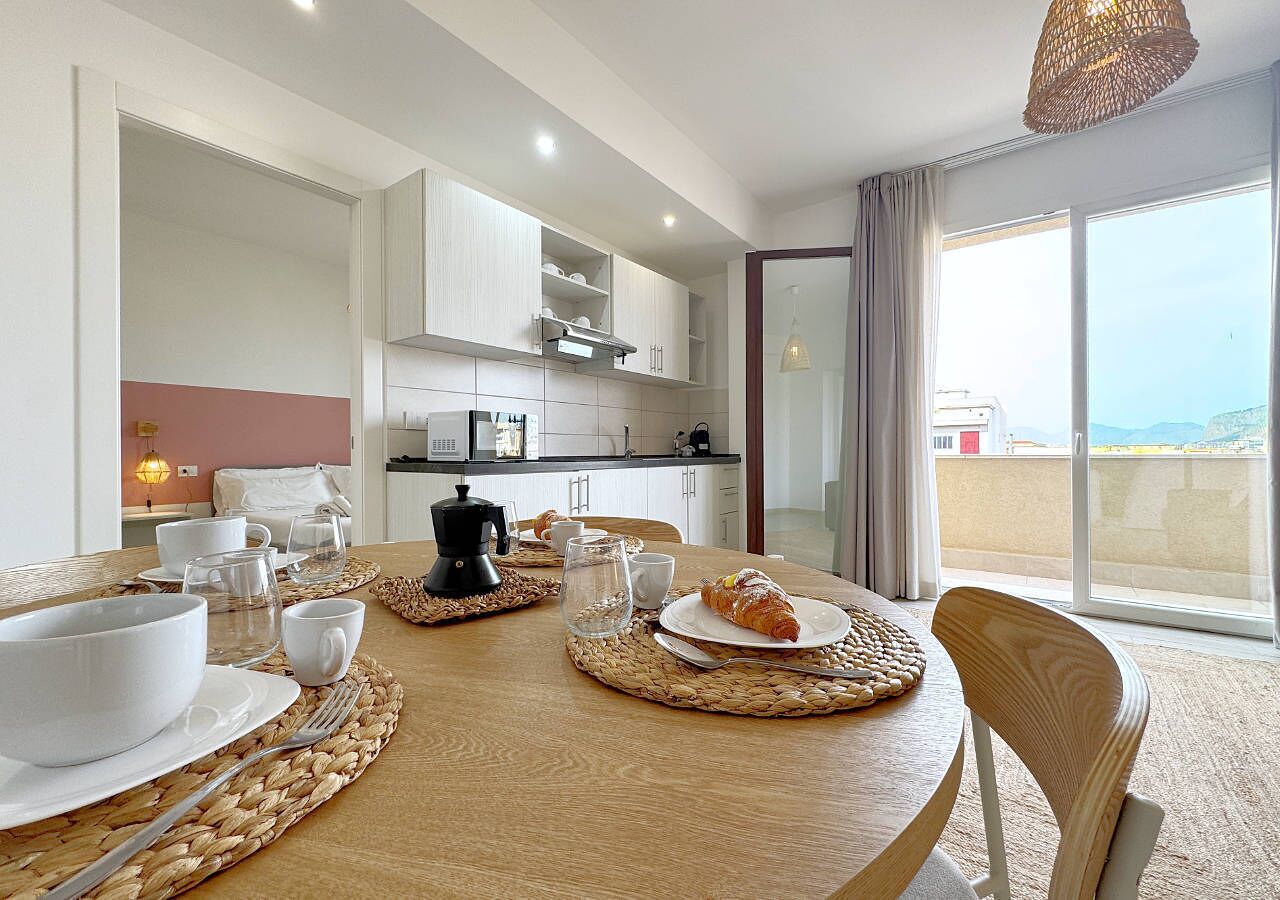 A dining area with a round table set for breakfast, a kitchen in the background, and a view of the sea through large windows.