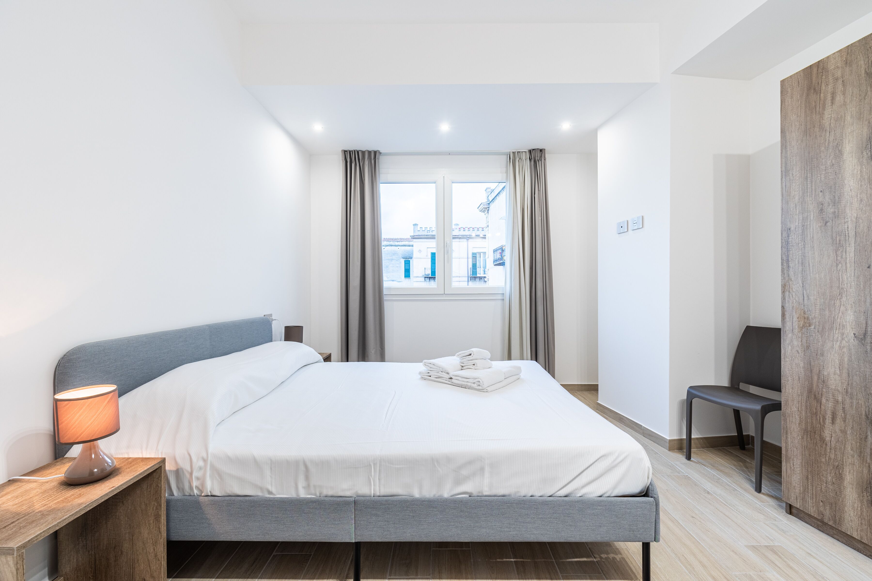 Een moderne hotelkamer met een bed, een bureau met lamp, een stoel en uitzicht op gebouwen door het raam.