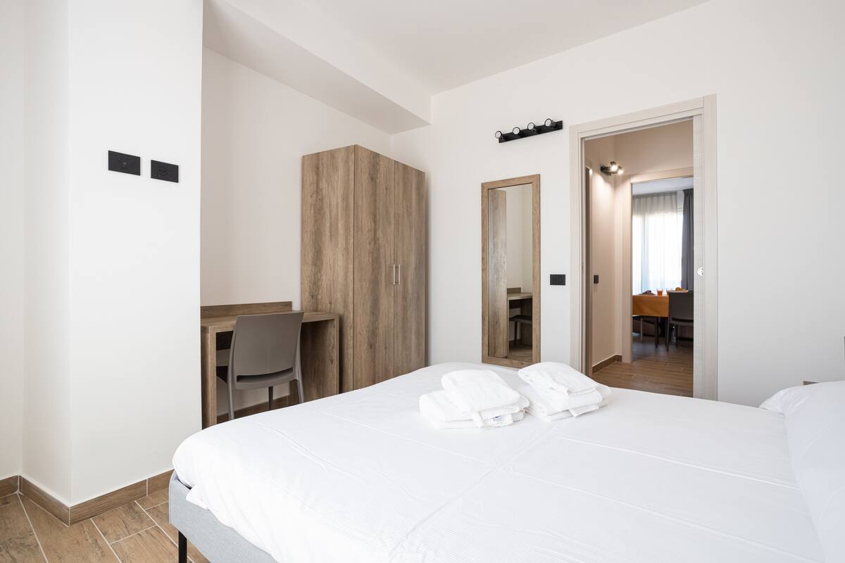 A hotel room with a large bed, a wooden wardrobe, a desk with a chair, a mirror, and a bathroom visible through an open door.