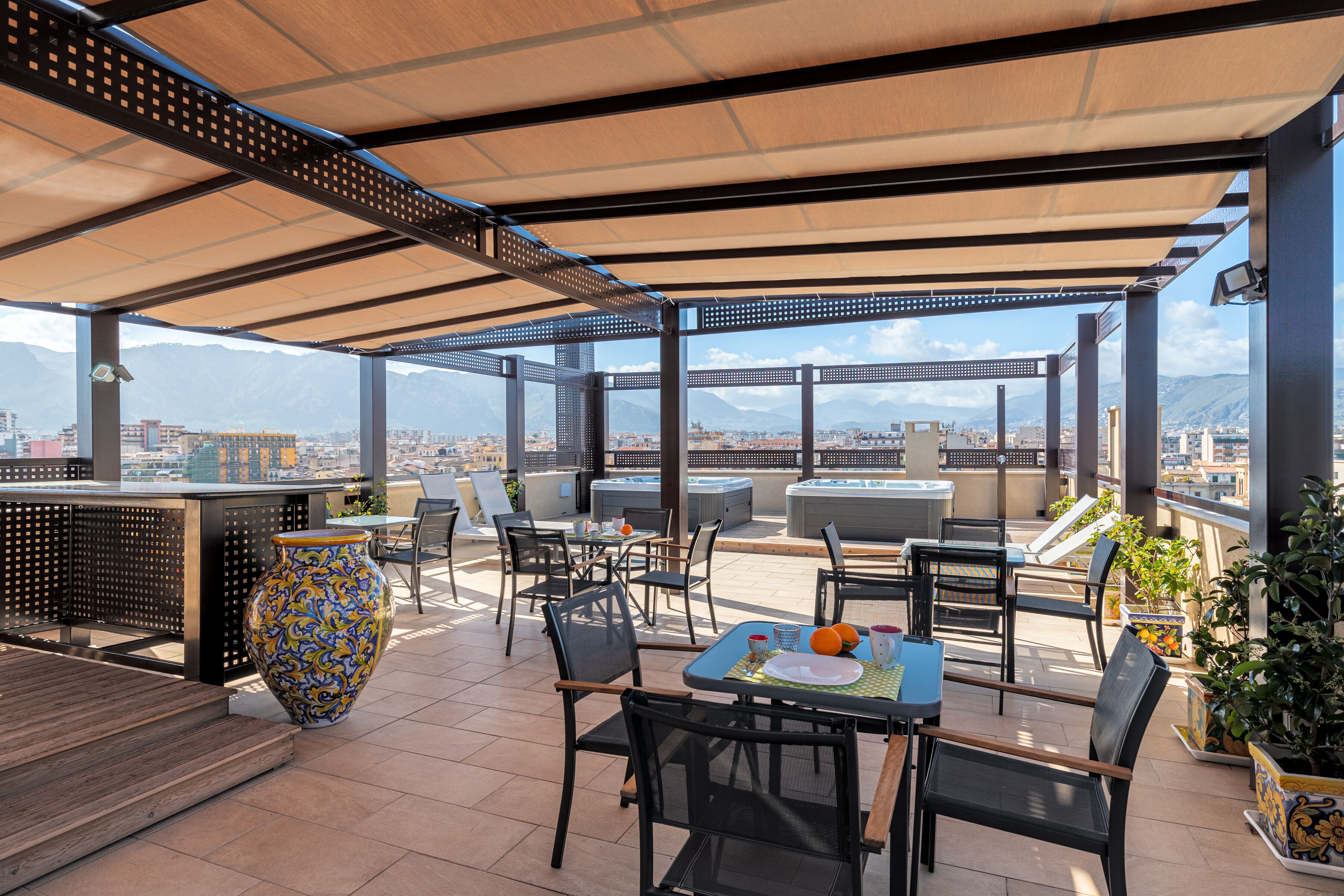 Une terrasse extérieure avec des tables et des chaises, un grand vase décoratif et une vue sur la ville et les montagnes.