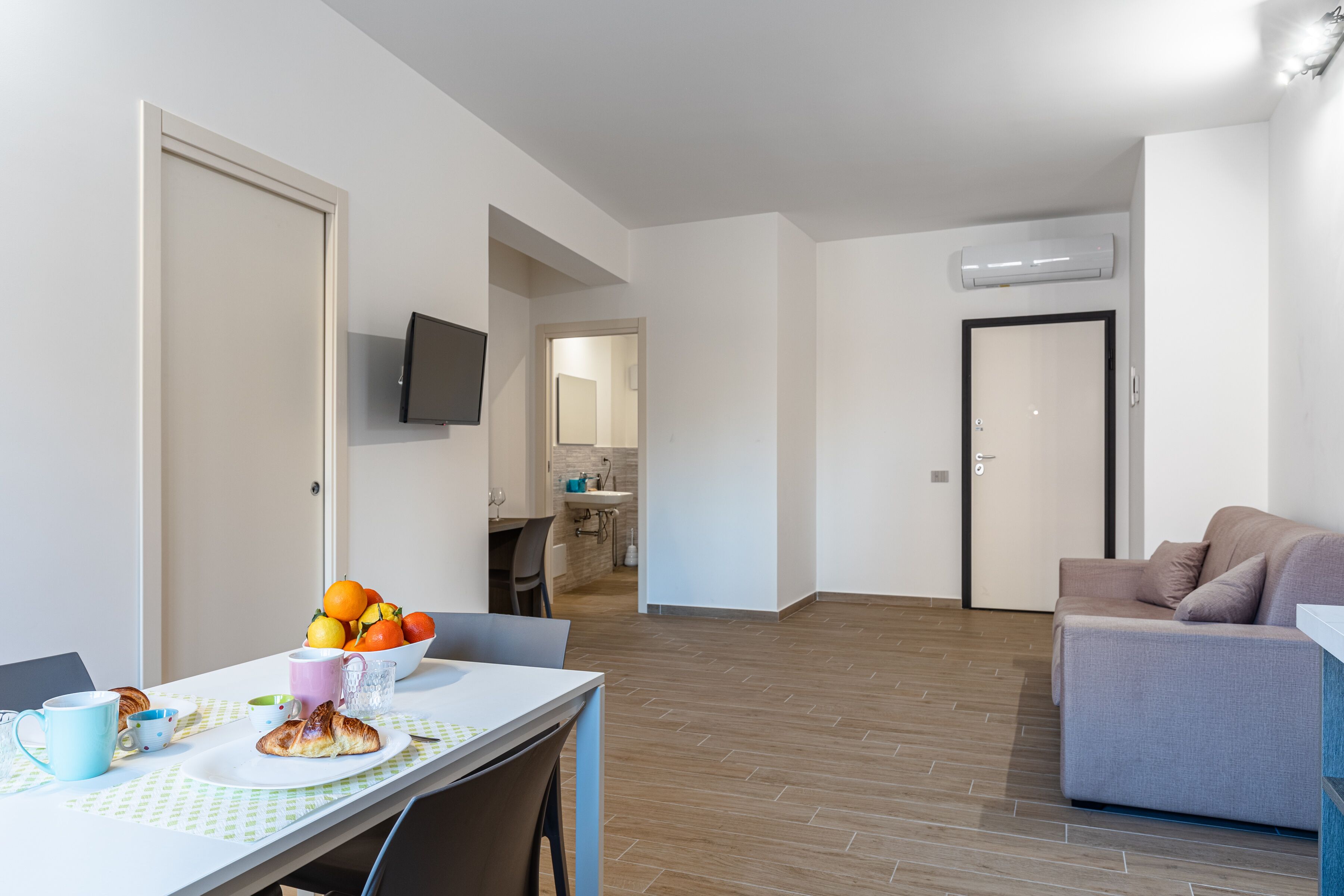 Una sala de estar moderna con un sofá gris, un comedor con una mesa dispuesta para el desayuno, y una cocina al fondo.