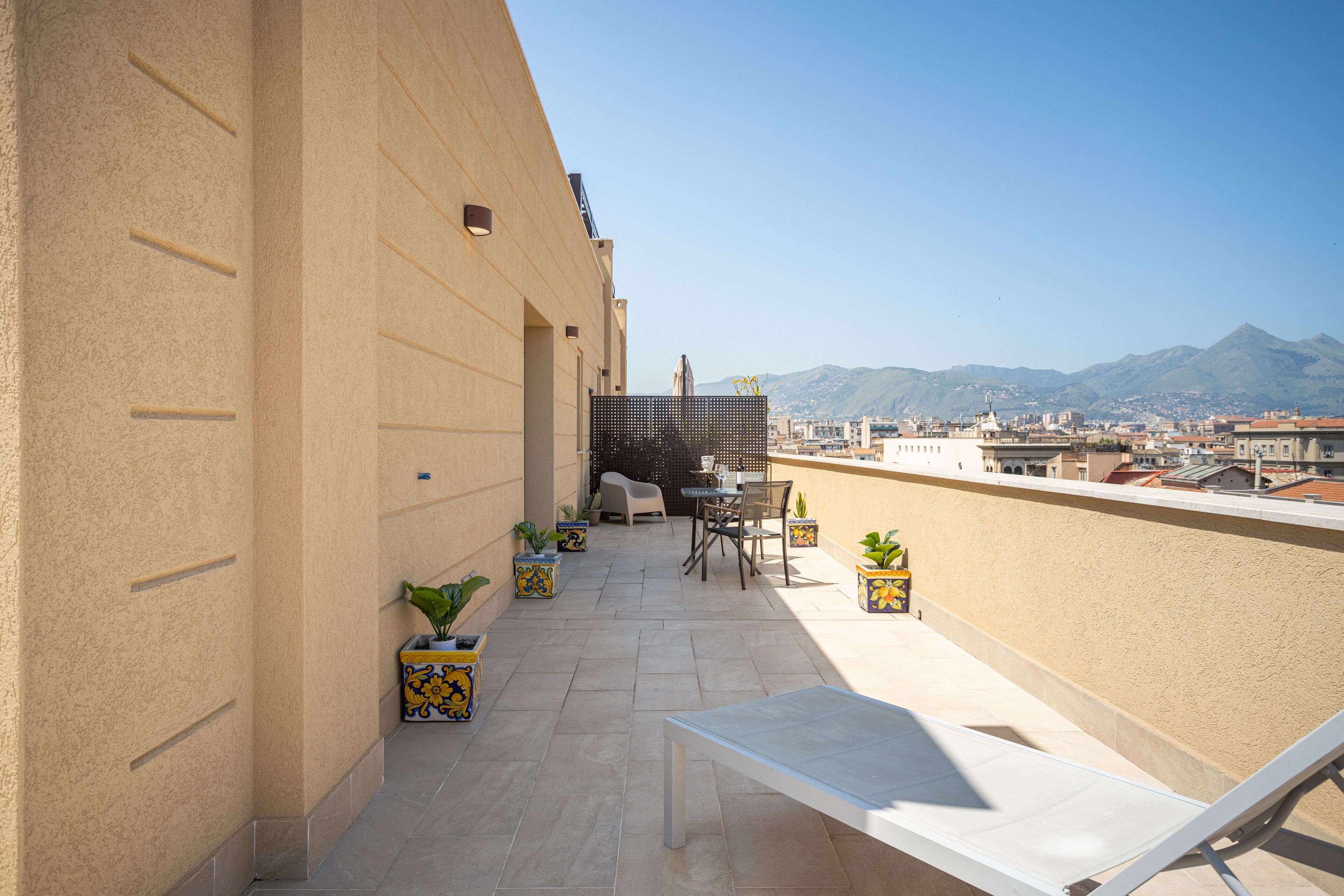 A rooftop terrace with a table and chairs, potted plants, and a view of the city and mountains.