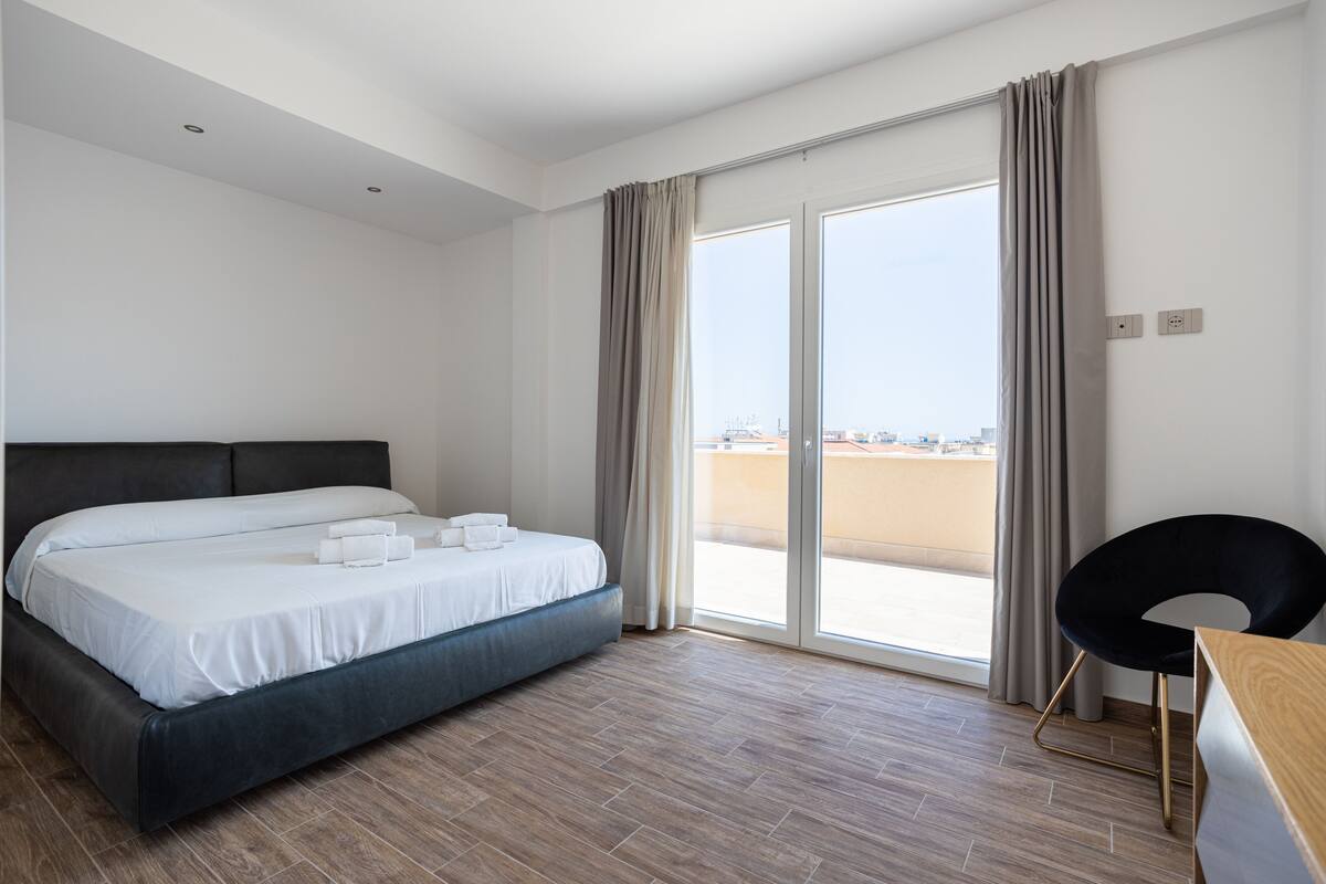 A modern hotel room with a large bed, a wooden desk, and a view of the beach through the sliding glass door.