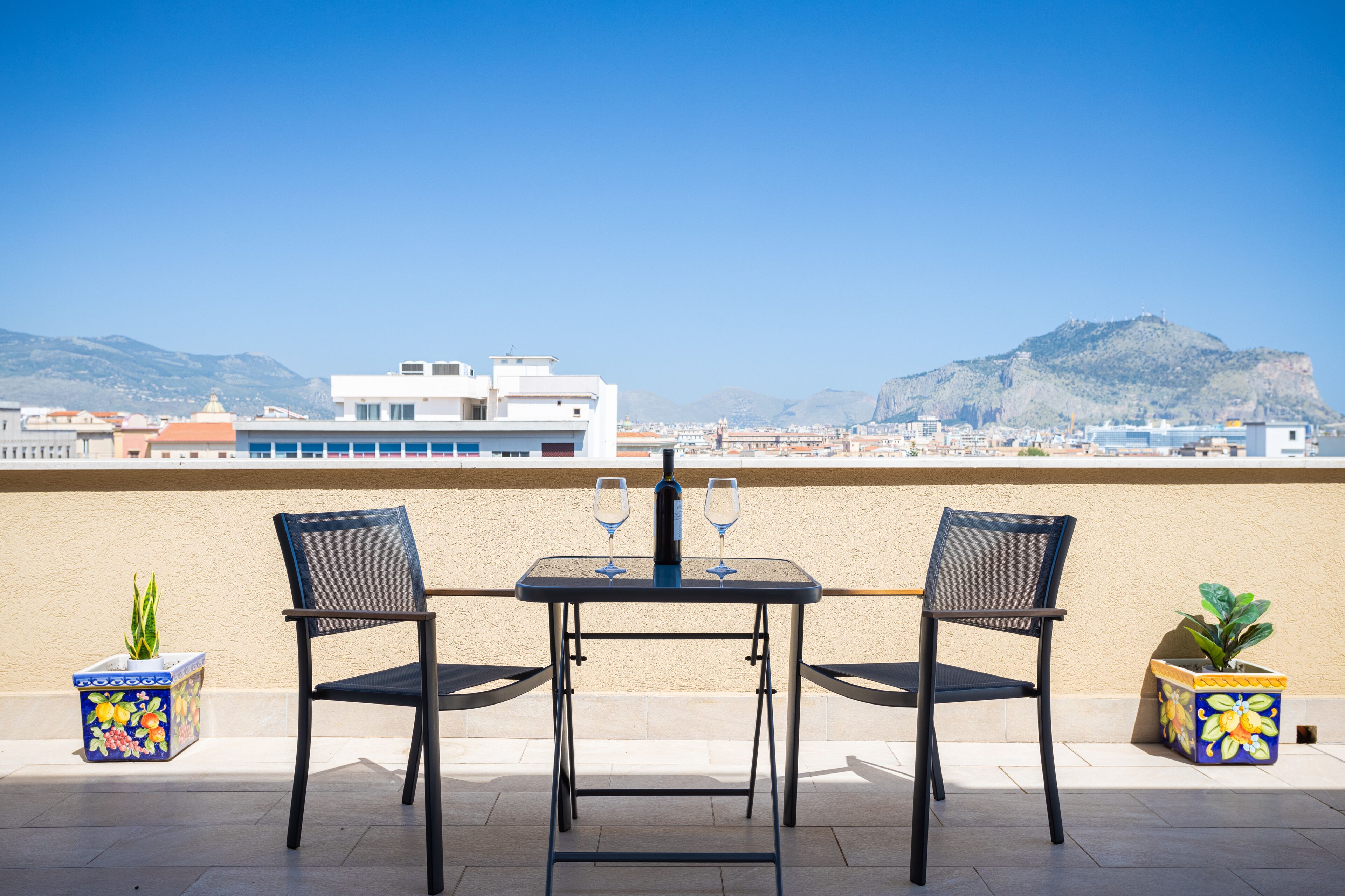 A terrace with a table and two chairs, a bottle, and two wine glasses, overlooking a cityscape with mountains in the background.