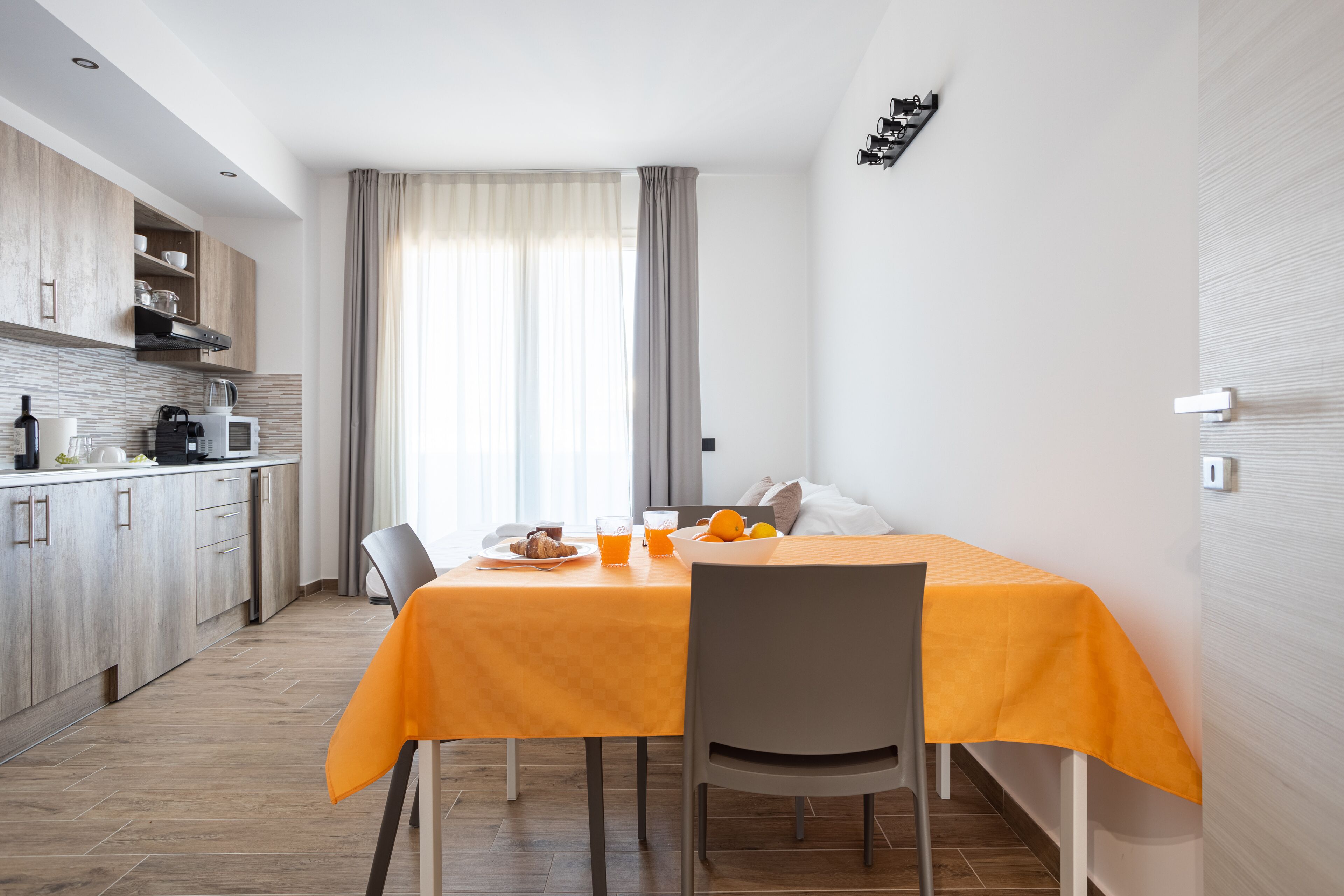 Une cuisine moderne avec des meubles en bois, une table à manger recouverte d’une nappe orange et un coin petit-déjeuner comprenant du jus d’orange et des toasts.