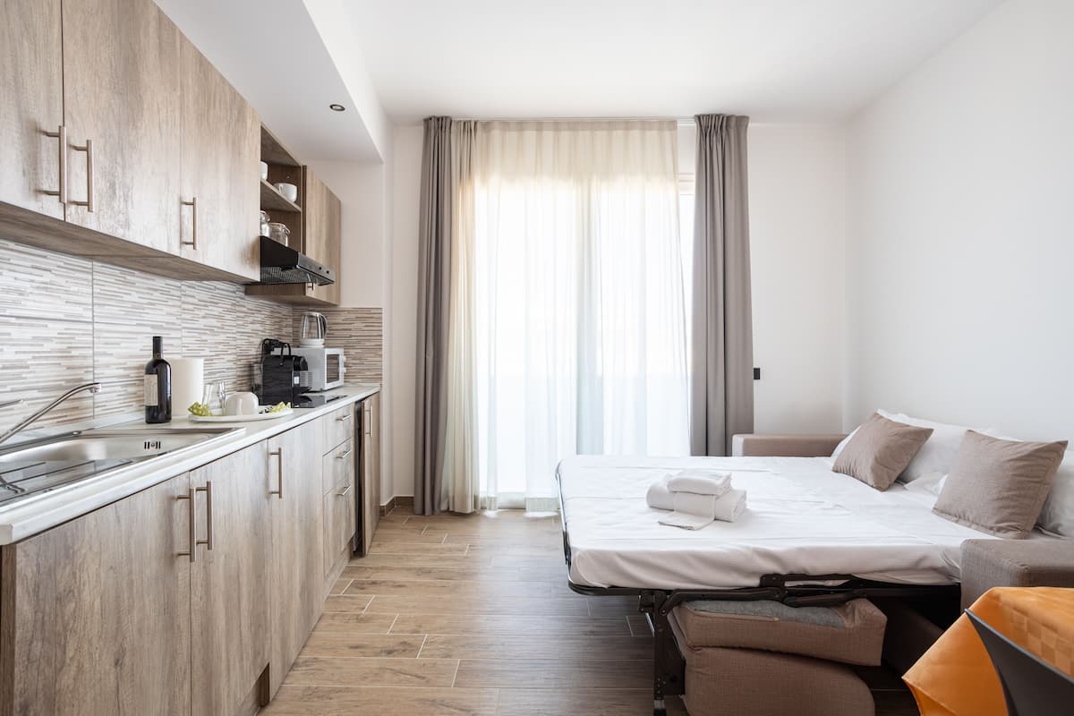 A modern hotel room with a kitchenette, a bed with pillows, and a large window with curtains.