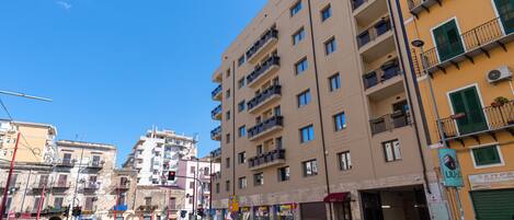 A multi-story building with balconies, a street with cars, and a clear blue sky.