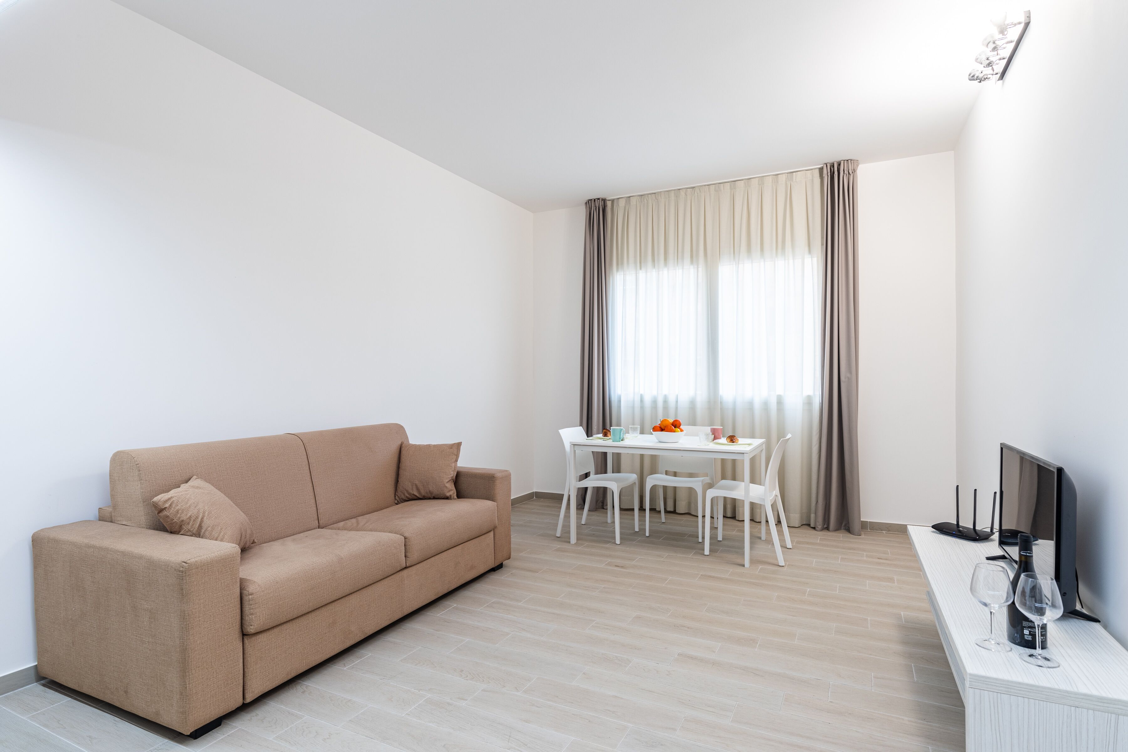 A modern living room with a beige sofa, a dining table with chairs, a flat-screen TV, and a large window with curtains.