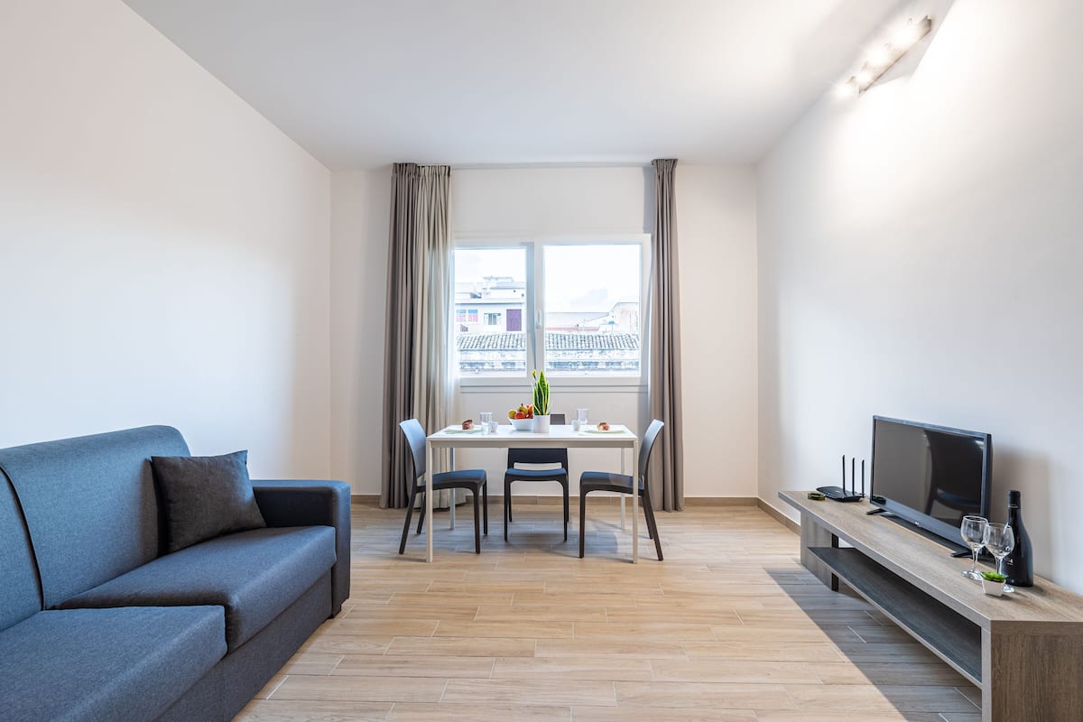 A modern living room with a sofa, dining table, chairs, a TV, and a window with curtains.