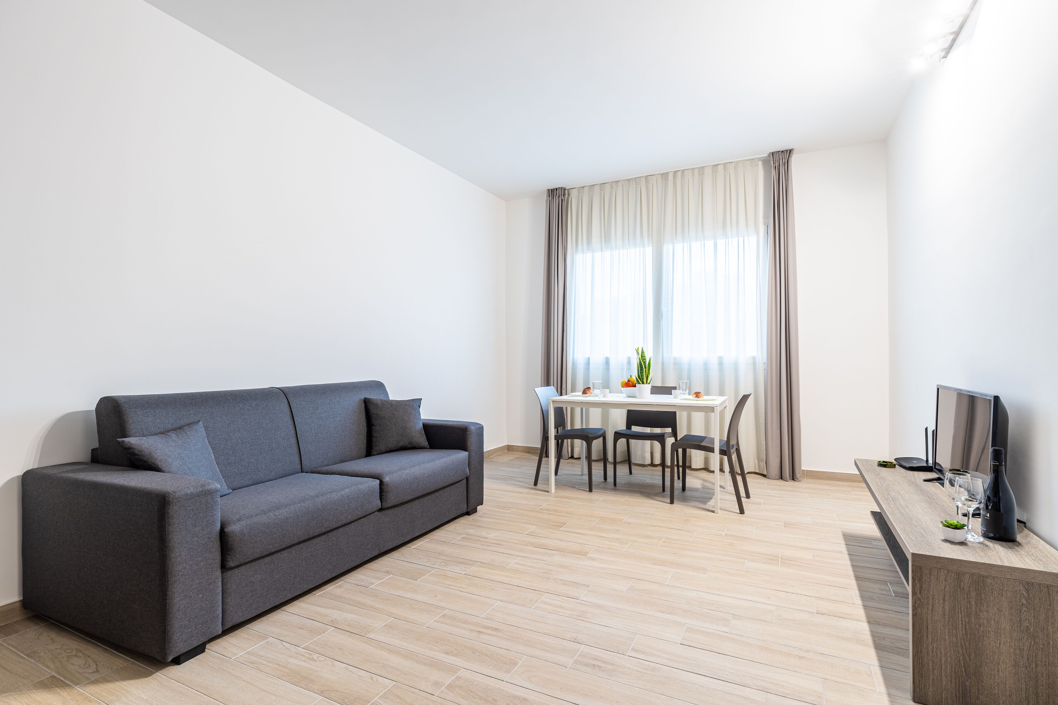 A modern living room with a grey sofa, a dining table with chairs, a TV on a wooden stand, and a large window with curtains.