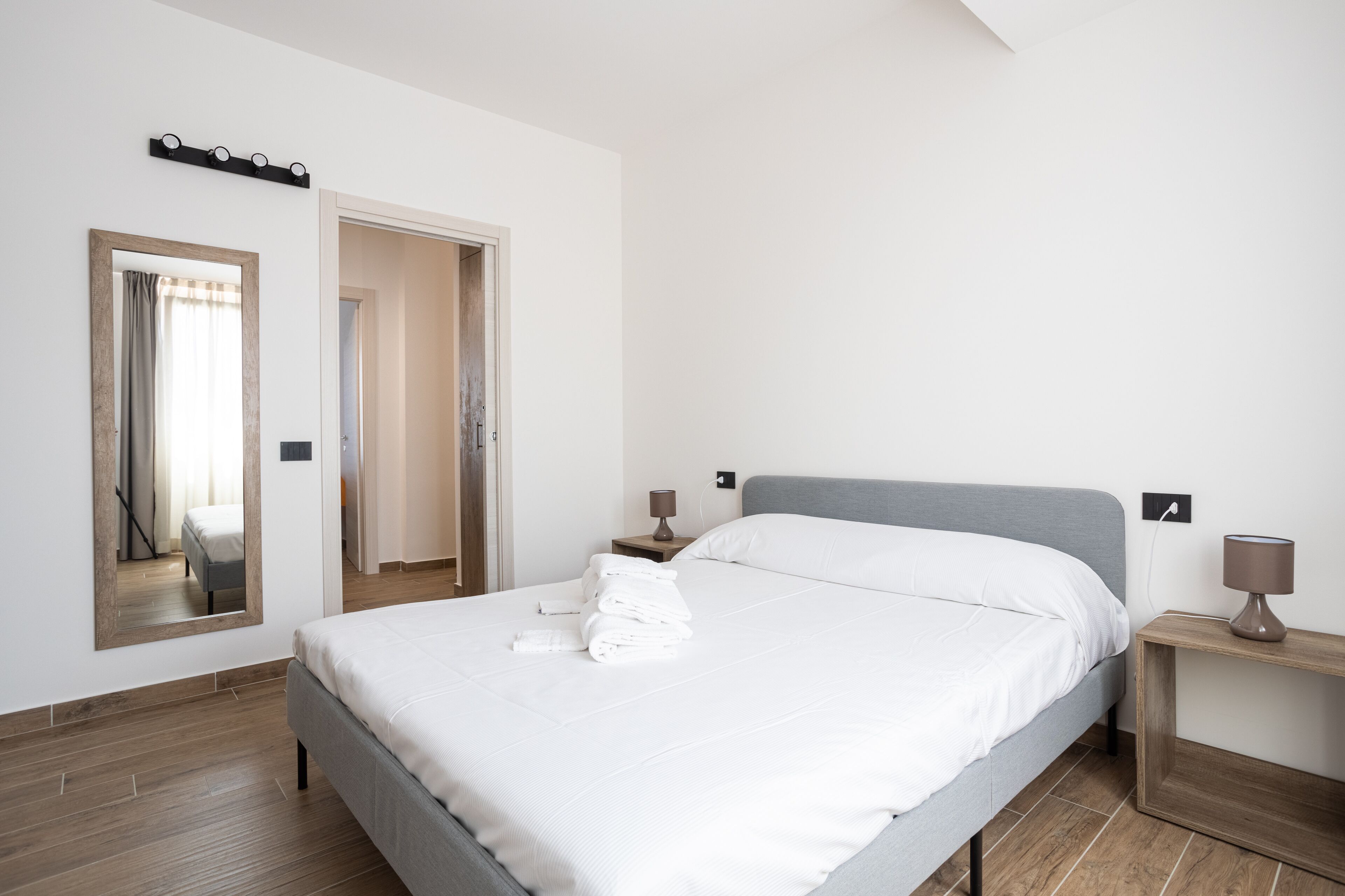 A neatly made bed with white linens and a grey headboard, two bedside tables with lamps, a large mirror, and a wooden floor.
