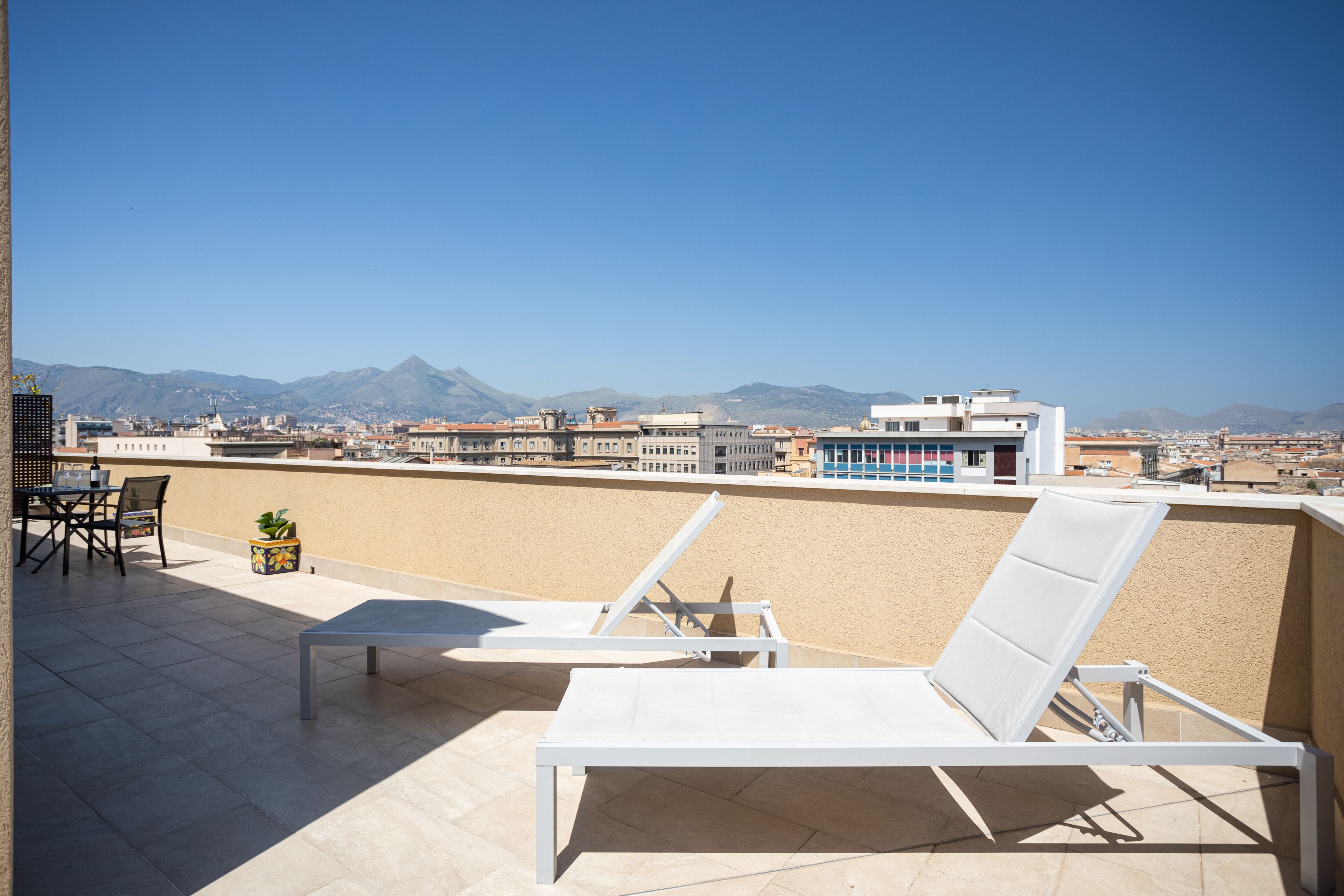 Eine Dachterrasse mit zwei weißen Liegestühlen, einem kleinen Tisch und einem Blumentopf. Von der Terrasse hat man einen Blick auf eine Stadtlandschaft mit Bergen im Hintergrund.