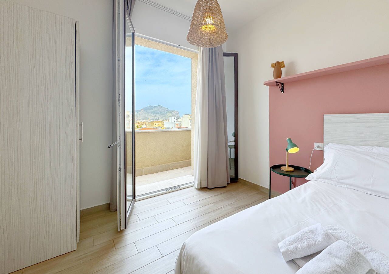 Un dormitorio con cama, mesita de noche, lámpara, ventana con cortinas y vista a la ciudad.