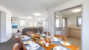Uma sala de estar moderna e bem iluminada, com uma mesa de jantar posta para uma refeição, um sofá e uma cozinha ao fundo.