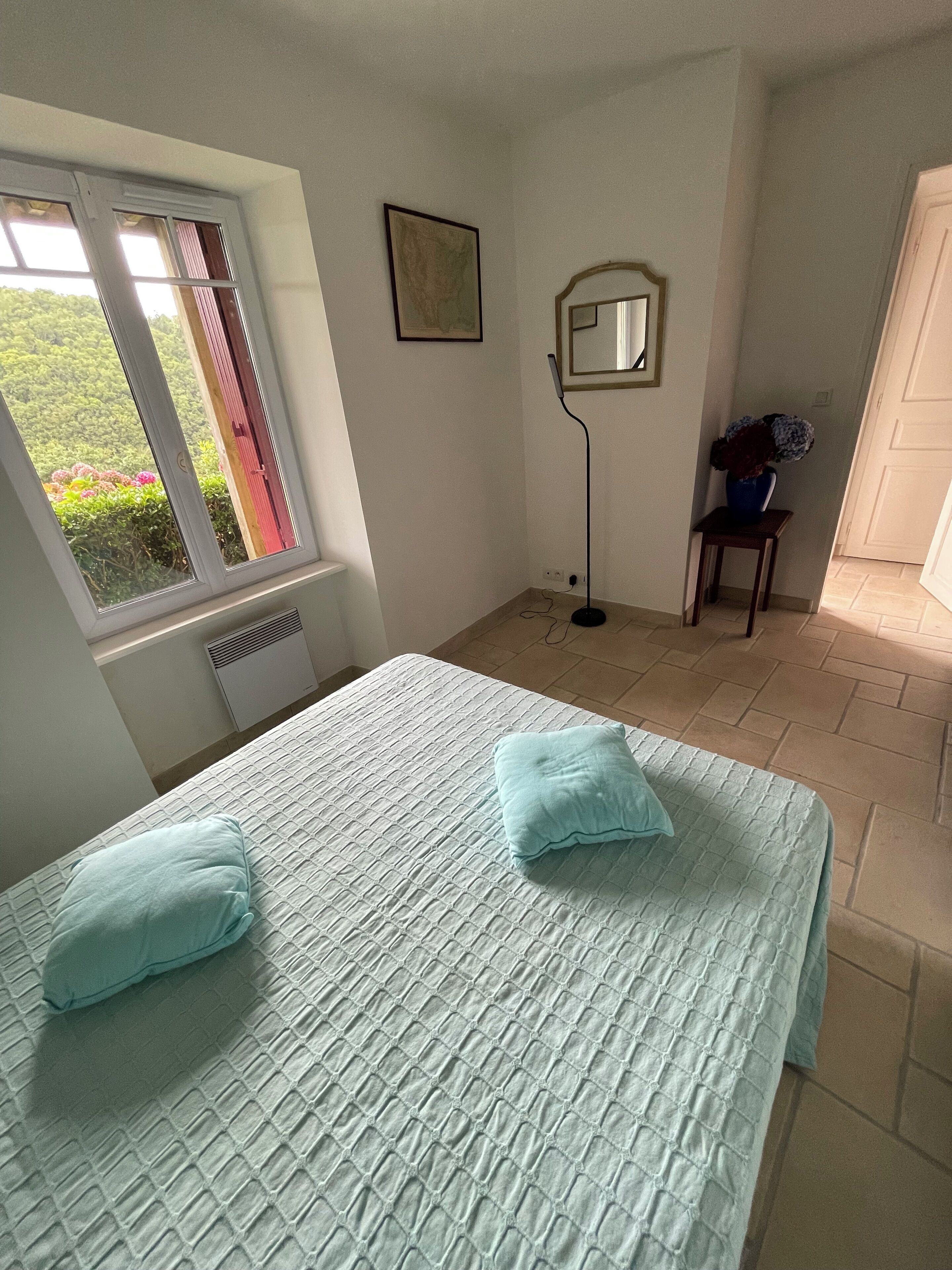 A bedroom with a bed, two pillows, a window, a standing lamp, a mirror, and a small table with a vase.