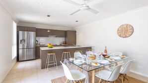Una cocina moderna con una isla central, electrodomésticos de acero inoxidable y una zona de comedor con mesa de vidrio y sillas blancas.