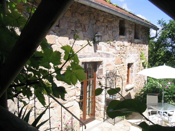 A stone building with a window and a door, a metal chair, a table, and an umbrella. - Charming stone house restored in English style (Arfeuilles)