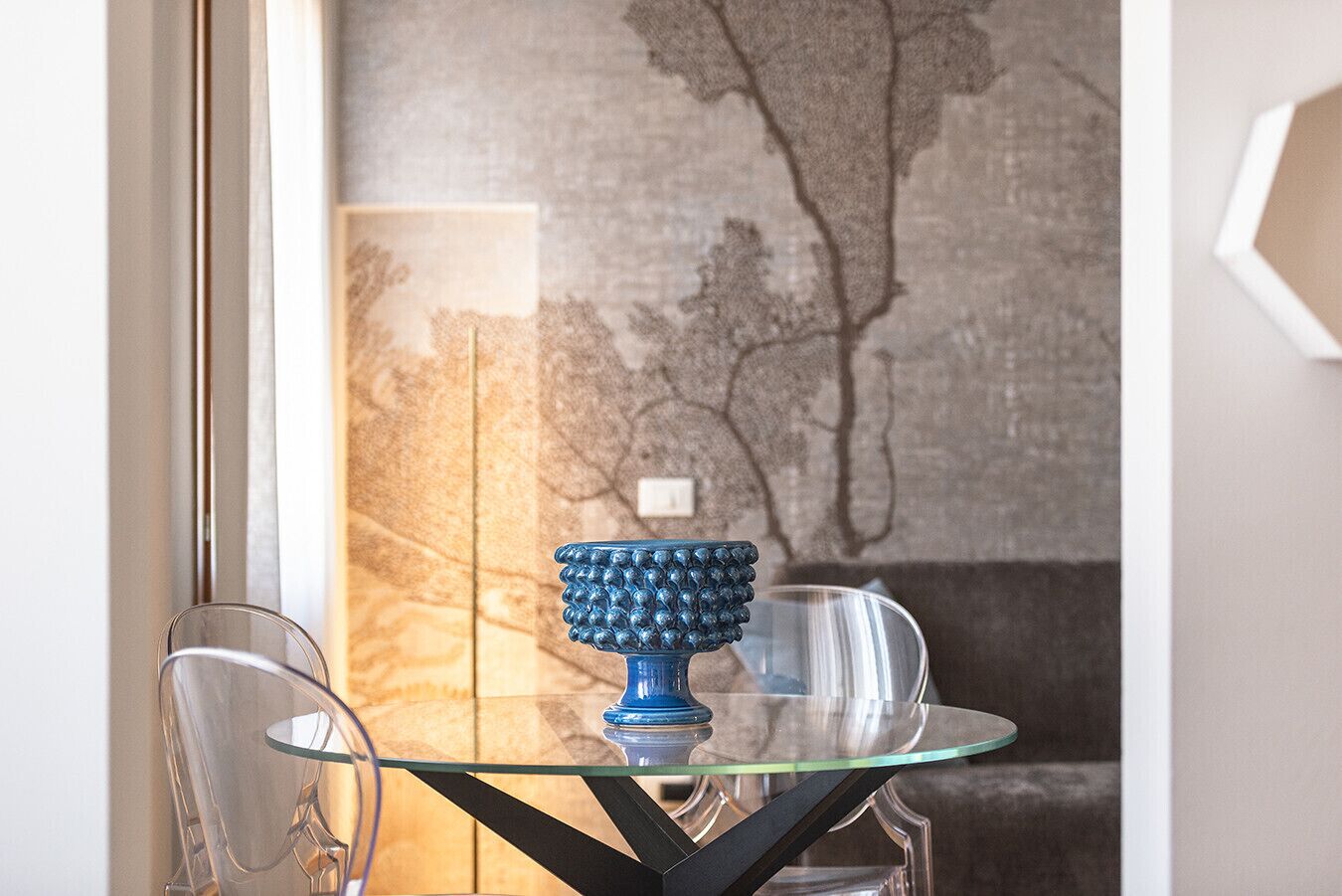 Una mesa de vidrio con un adorno azul y sillas transparentes en una habitación con pared texturizada y ventana.