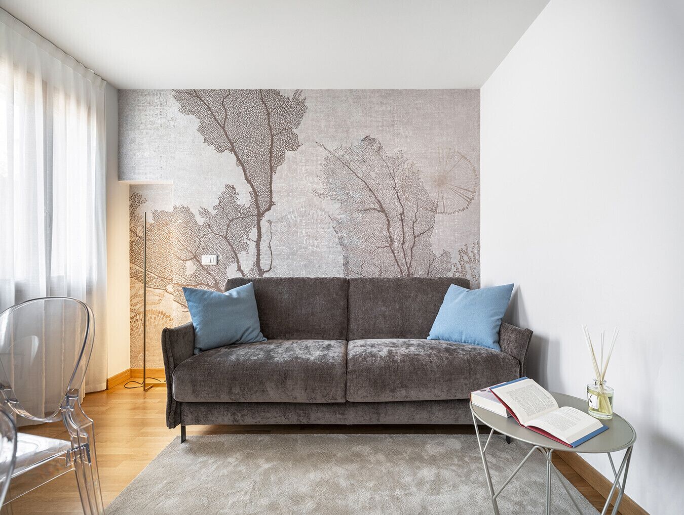 Una sala de estar moderna con un sofá gris, dos cojines azules, una silla de vidrio transparente, una mesita redonda con un libro y un difusor de aroma de caña, y un gran mural abstracto con la imagen de un árbol en la pared.