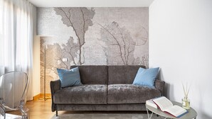 Un soggiorno moderno con un divano grigio, due cuscini blu, una sedia trasparente in vetro, un tavolino rotondo con un libro e un diffusore di fragranze, e un grande murale astratto raffigurante un albero.