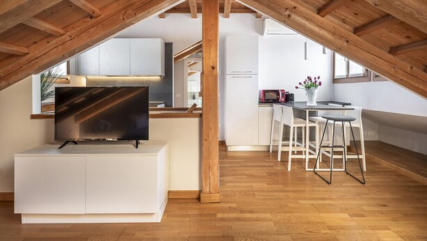 Ein modernes Loft mit freiliegenden Holzbalken, einem Flachbildfernseher, einem Essbereich mit weißem Tisch und Stühlen sowie einer Küche mit weißen Schränken.