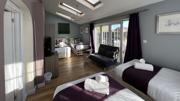 A modern bedroom with two beds, a sofa, a TV, and a large window with natural light. - Lancaster Close Serviced Accommodation (Bicester)