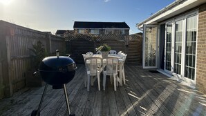 A wooden deck with a barbecue grill, a dining table with chairs, and a brick building with large windows. - Lancaster Close Serviced Accommodation (Bicester)