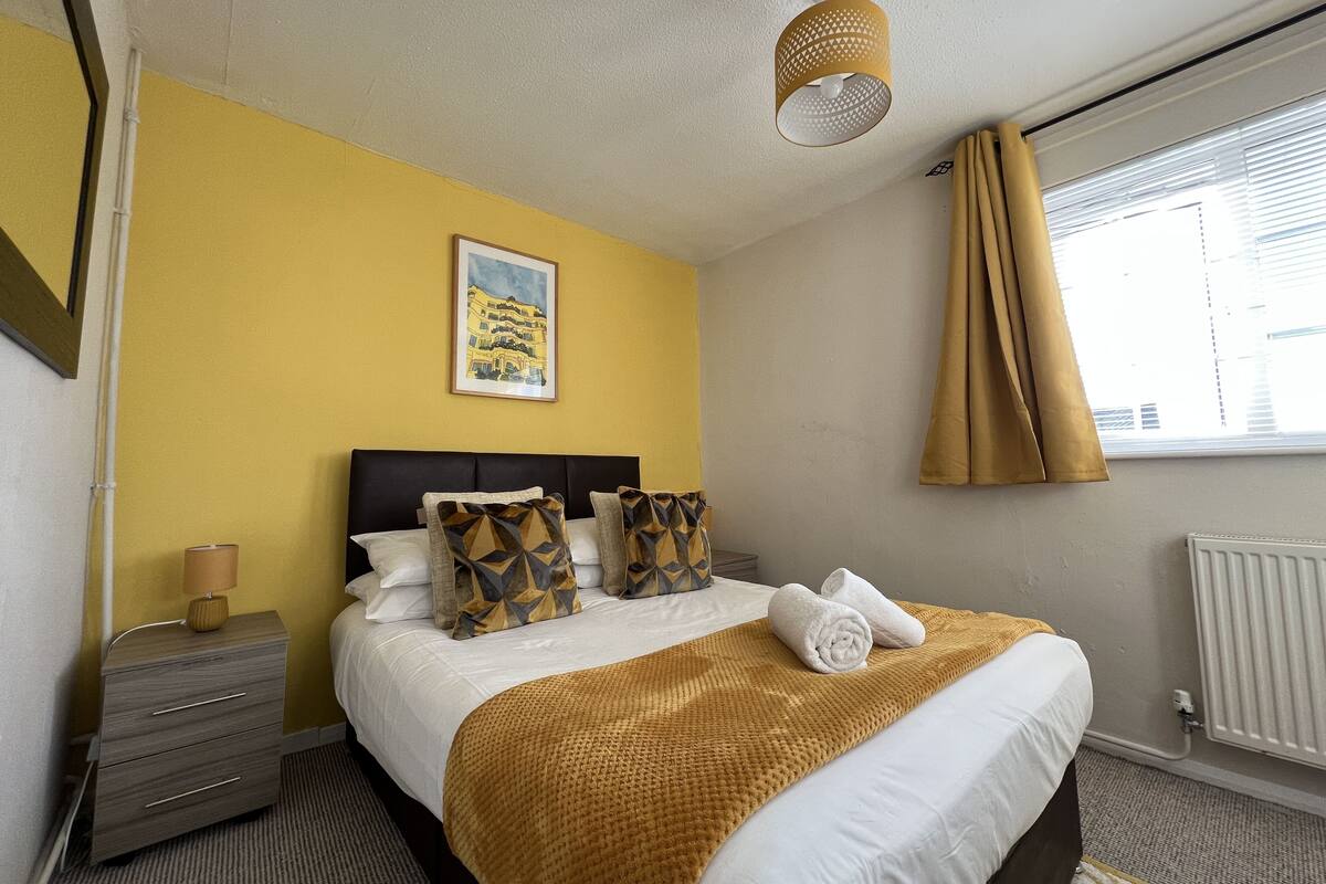 Una habitación de hotel con cama, mesita de noche, lámpara, un cuadro y ventana con cortinas.