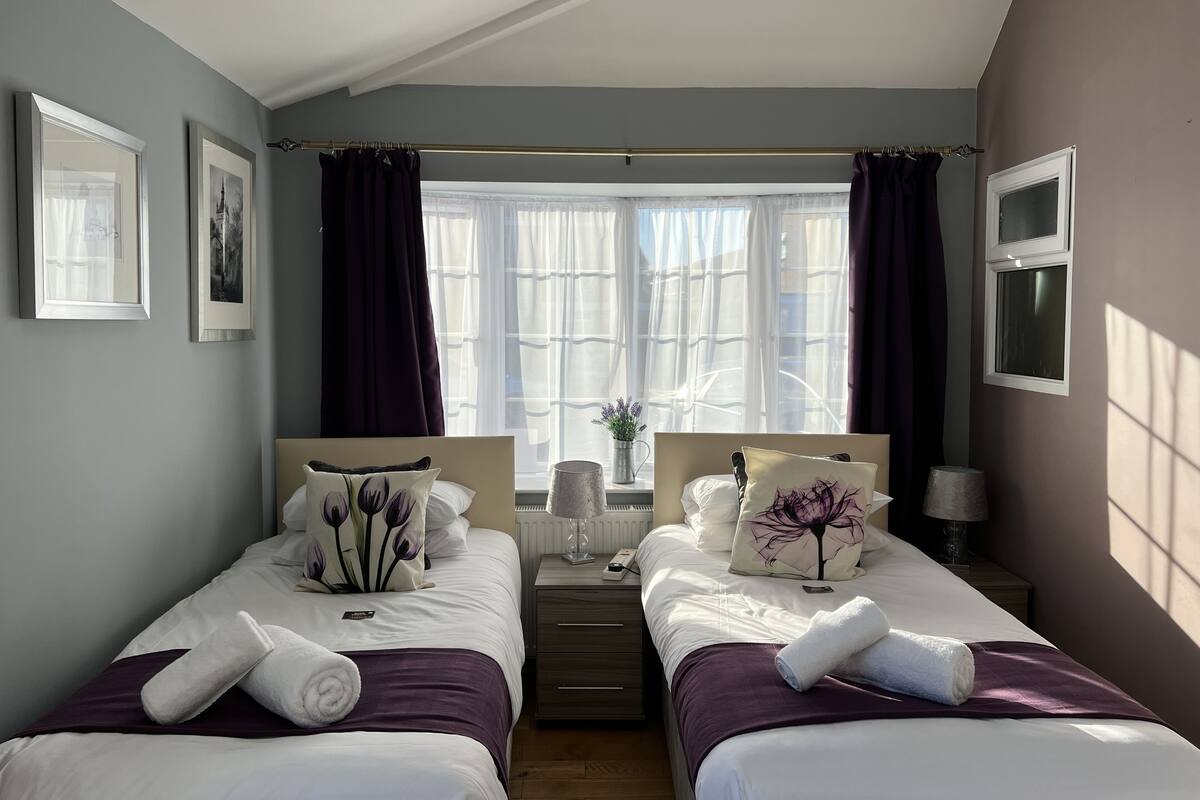 Habitación de hotel con dos camas, una ventana con cortinas translúcidas, una mesita con una lámpara y cuadros en las paredes.