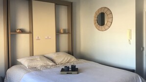 A neatly made bed with a tray of two black cups, a wall-mounted mirror, and a bedside table with a lamp.