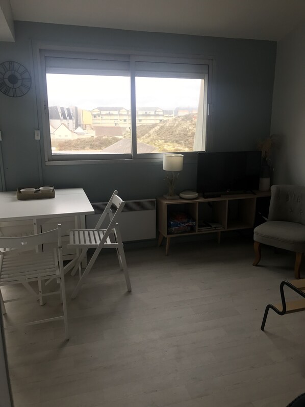 A room with a large window, a dining table with chairs, a TV, and a lamp. - Studio Le Sandettie, 50 meters from the beach! + private garage (Fort-Mahon-Plage)